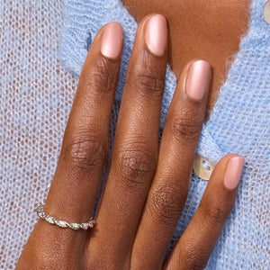A hand with light pink polished nails holds a bottle of MANUCURIST - GREEN™ Nail Polish in Carnation by MANUCURIST INC, featuring a pearly pink vegan formula and the “green” label, resting against the pocket of light blue denim jeans.