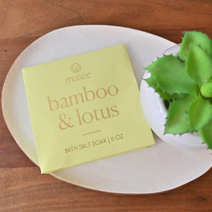 A MUSEE - Bamboo & Lotus Mini Bath Salt Soak from MUSEE rests on a white plate beside a green potted succulent, all arranged atop a wooden surface.