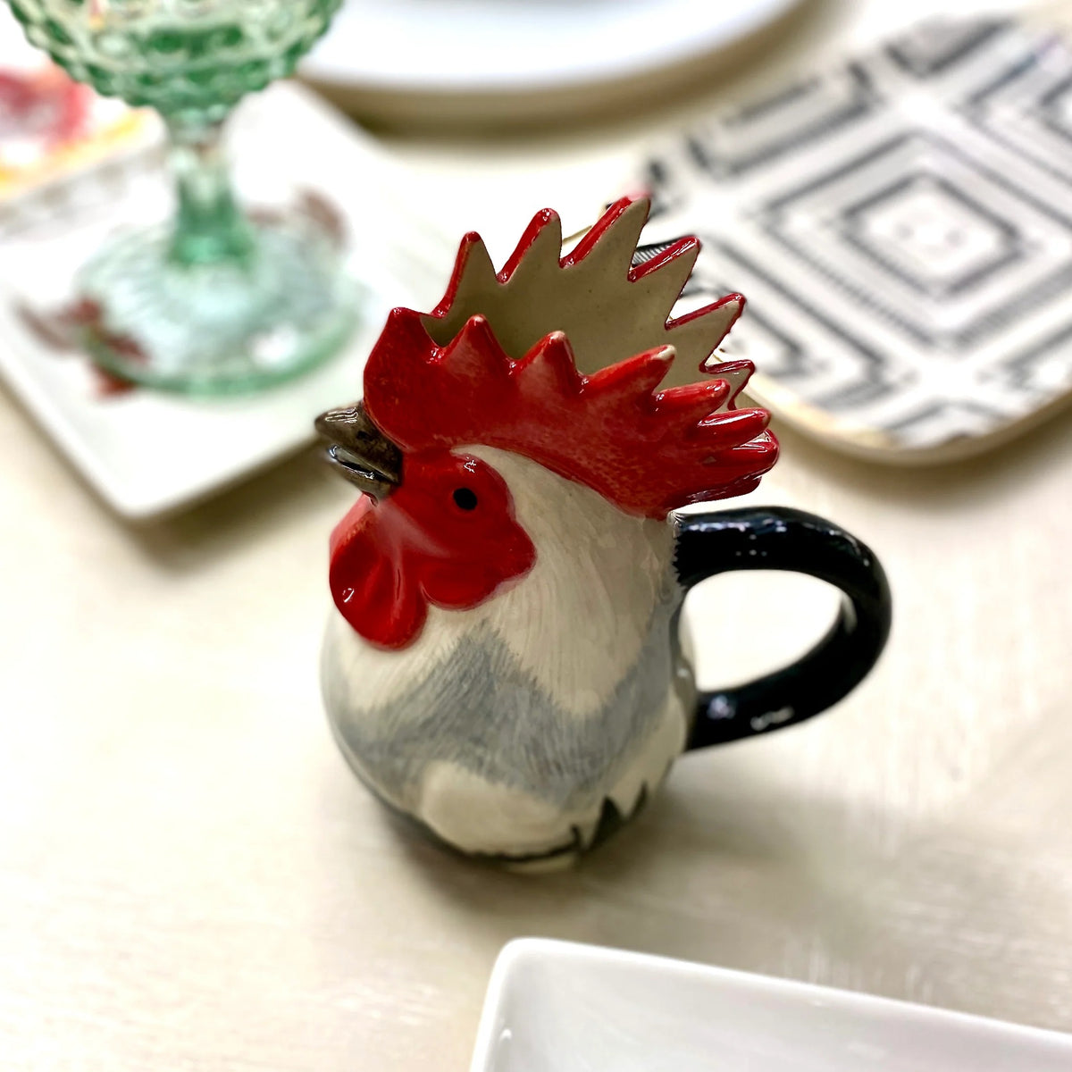 The QUAIL CERAMICS - DORKING ROOSTER JUG by QUAIL DESIGNS, a hand-painted stoneware jug with a red comb and black handle, sits on the table beside a green glass and patterned plates—a charming British tea accessory.