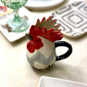 The QUAIL CERAMICS - DORKING ROOSTER JUG by QUAIL DESIGNS, a hand-painted stoneware jug with a red comb and black handle, sits on the table beside a green glass and patterned plates—a charming British tea accessory.