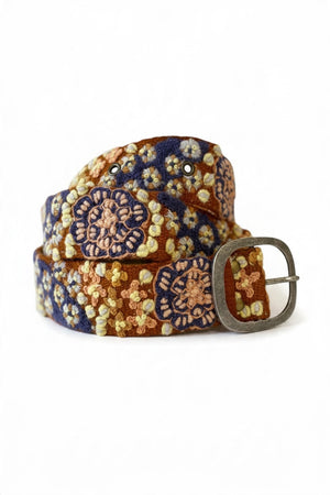 A close-up of a person in a blue denim shirt, dark pants, and the JENNY KRAUSS BLUE SKIES BELT, featuring vibrant floral embroidery and a large oval buckle—handcrafted in Peru.