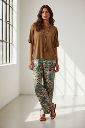 A woman with long brown hair wears the MOMONI - IORA LUREX JERSEY T SHIRT in gold, paired with beige and blue wide-leg floral pants and brown sneakers, standing against a neutral studio background.
