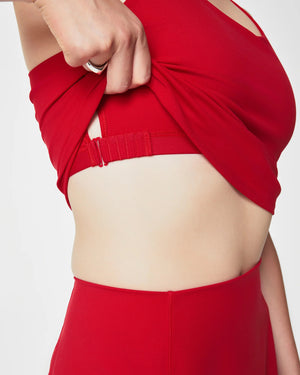 A woman with hands on hips smiles and looks to the side, wearing the SPANX - CONTOUR CROSSBACK TANK IN SPANX RED, a sleeveless athletic top with a flared hem and built-in shelf bra, against a plain white background.