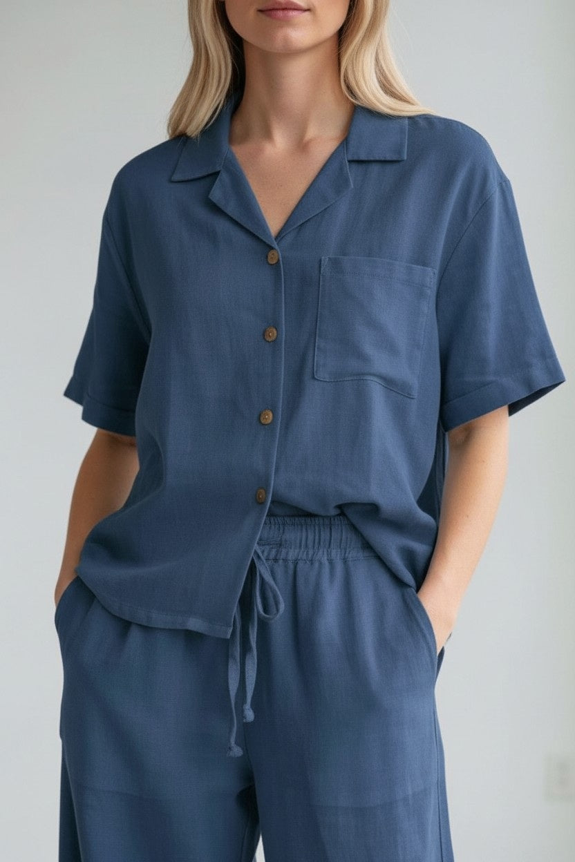 Wearing the LLEA BLUE - CASEY Short Sleeve Linen Shirt in Midnight Ocean, styled with matching drawstring blue pants, this individual stands with hands in pockets. The relaxed silhouette and lightweight linen fabric from LLEA BLUE make it ideal for warm-weather days. Face is partially out of frame.