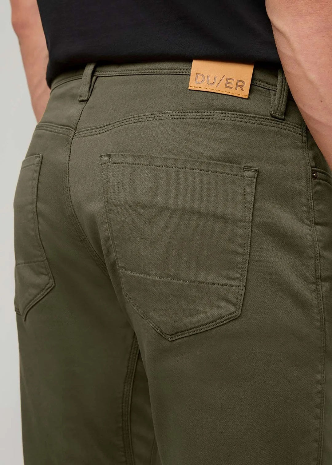 A person stands facing away from the camera against a plain background, wearing DUER - NO SWEAT ATHLETIC TAPER PANTS IN KELP paired with brown shoes.