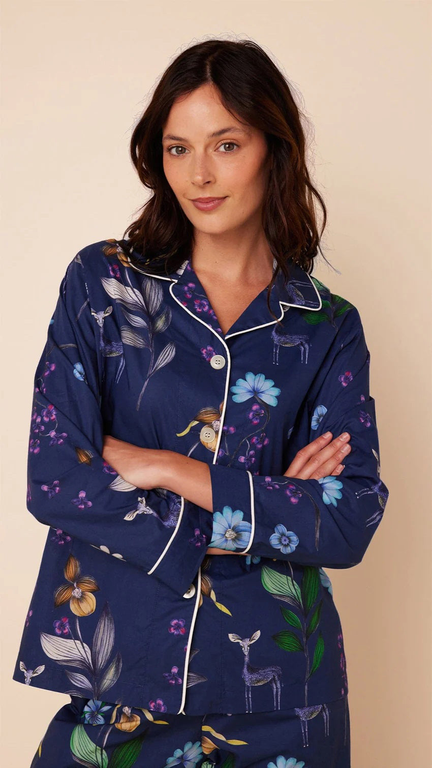 A woman stands barefoot against a plain background, wearing the CAT&#39;S PAJAMAS - Deerly Luxe Pima Cotton Pajamas by CAT&#39;S PAJAMAS. The navy blue pajama set features a colorful floral pattern and is crafted from ultra-soft pima cotton. It includes a long-sleeved button-up shirt and matching pants, both adorned with white piping along the edges.