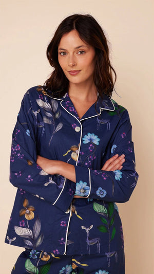 A woman stands barefoot against a plain background, wearing the CAT'S PAJAMAS - Deerly Luxe Pima Cotton Pajamas by CAT'S PAJAMAS. The navy blue pajama set features a colorful floral pattern and is crafted from ultra-soft pima cotton. It includes a long-sleeved button-up shirt and matching pants, both adorned with white piping along the edges.