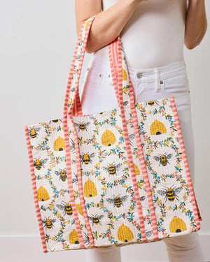 The PRINTFRESH - SWEET BEE TRAVELER TOTE from the Sweet Bee collection showcases floral and bee designs with yellow beehives on white, complemented by pink and white striped handles and borders for a charming contrast against a light beige wall.