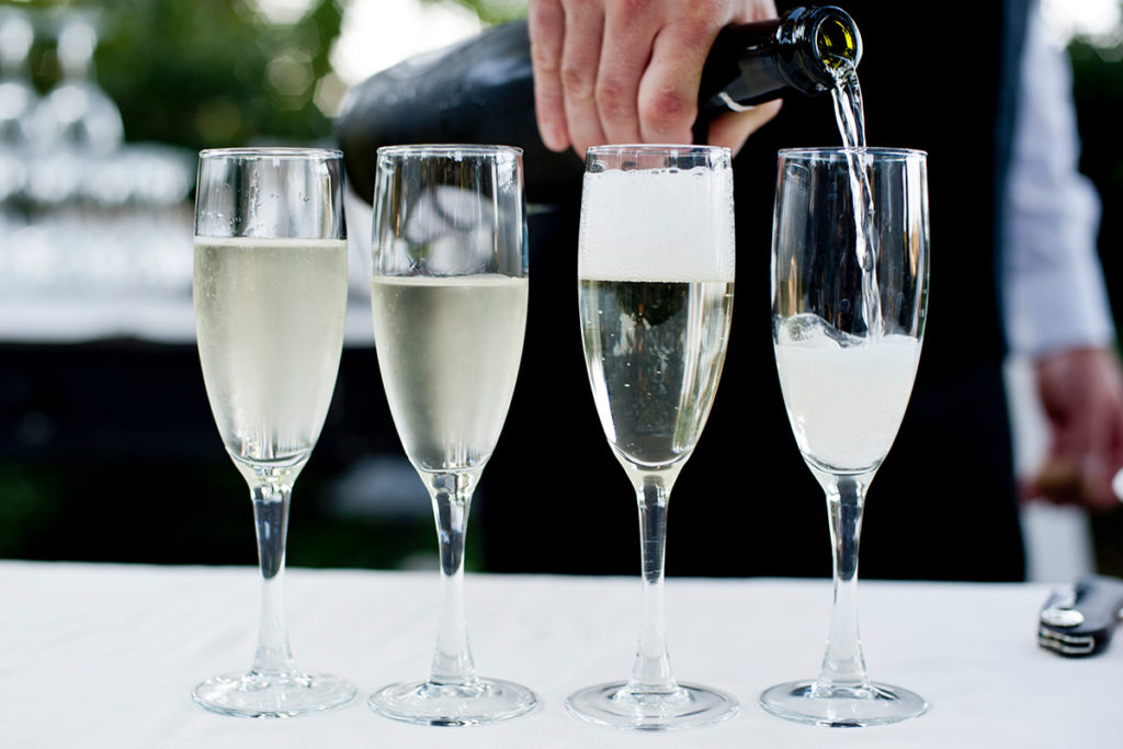 A person pours sparkling wine or champagne into one of four tall, clear glasses placed in a row on a white surface. The outdoor background is blurred.