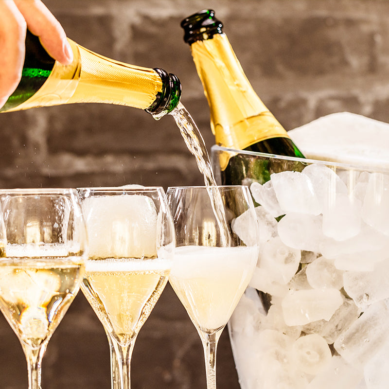 A hand pours champagne into three glasses, with two bottles—one being poured and one in an ice bucket—against a blurred brick background.