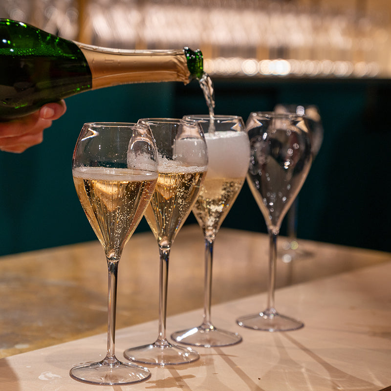 A hand pours sparkling wine or champagne into four elegant glasses on a wooden surface, with bubbles rising and foam forming as the glasses fill. The background is softly blurred.