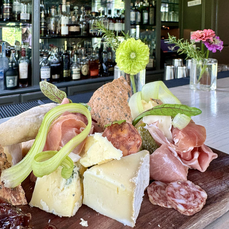 A close-up of a charcuterie board with assorted cheeses, cured meats, bread, and pickled vegetables, set on a bar counter with bottles of liquor and flowers in the background.