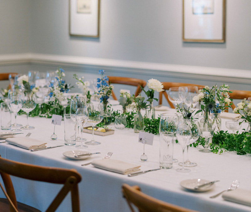 Elegant dining table set for an event, with white tablecloth, glassware, place cards, and green and white floral centerpieces. Wooden chairs are arranged around the table in a softly lit room.