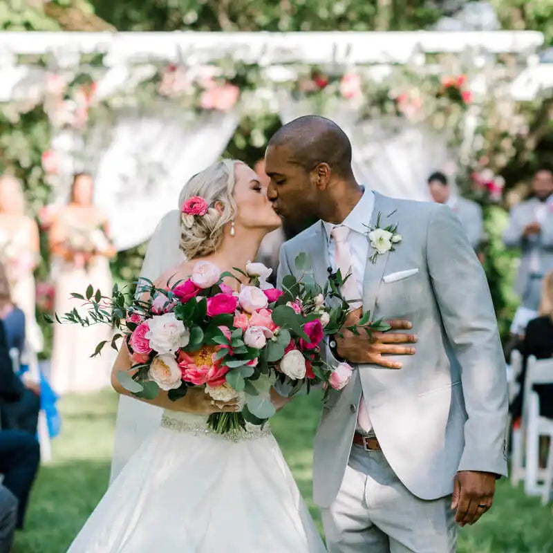 A bride and groom kiss outdoors at their wedding ceremony, holding a lush bouquet of pink and white flowers, with guests and floral decorations visible in the background.