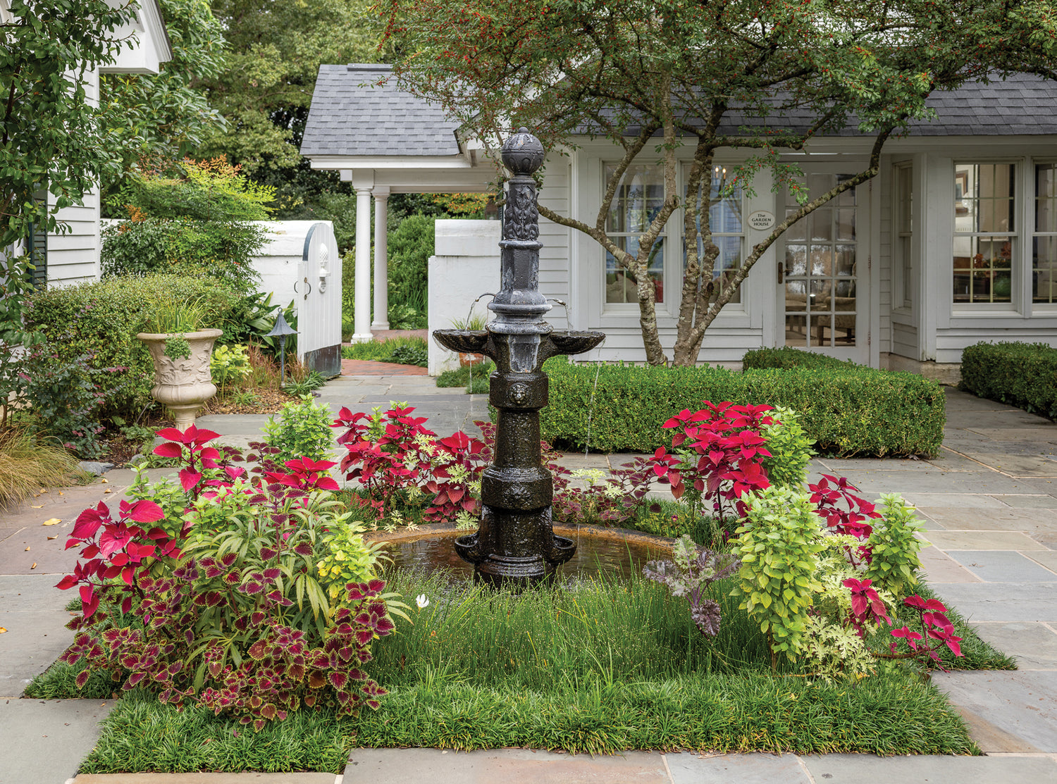 A black tiered fountain stands in the center of a small square garden bed bordered by green grass and colorful red and green foliage, set in front of a white house with trimmed hedges and paved stone paths.
