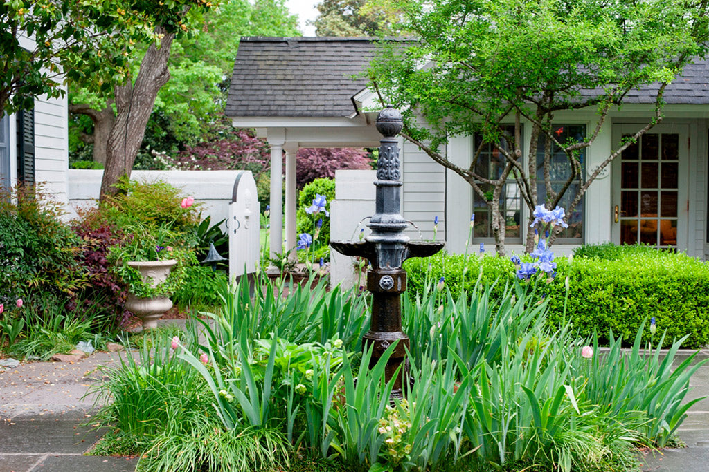 A black decorative garden fountain stands amid tall green plants and blooming flowers, surrounded by a paved walkway in front of a house with white siding and large windows.
