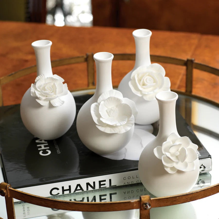 The CAMEO LONG NECK PORCELAIN BUD VASE by ZODAX is a round, white vase with a matte finish, featuring a narrow neck and a detailed floral embellishment on the front.