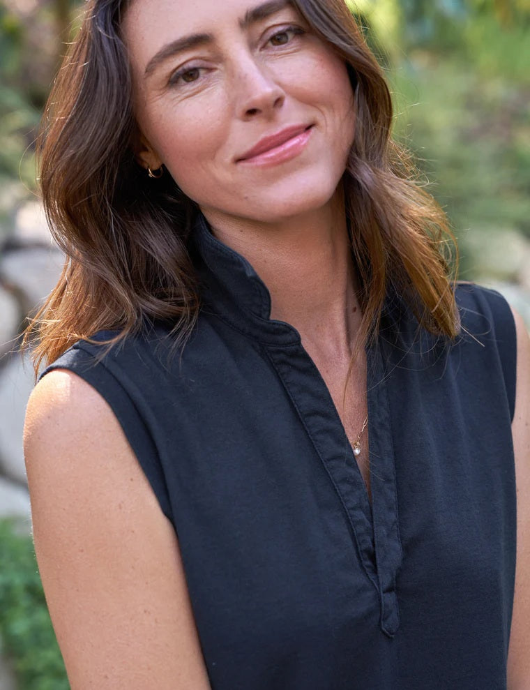 A woman stands outdoors, smiling and holding a woven basket of daisies, wearing the FRANK & EILEEN MINI LILY POPOVER HENLEY TANK DRESS in Vintage Jersey British Royal Navy. Lush greenery and a stone wall form the backdrop to this California-made look.