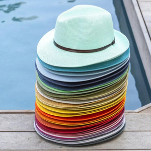 A tall stack of colorful, wide-brimmed hats is placed on a wooden deck next to a swimming pool. The hats are arranged in a gradient from light blues and greens at the top to reds and purples at the bottom.