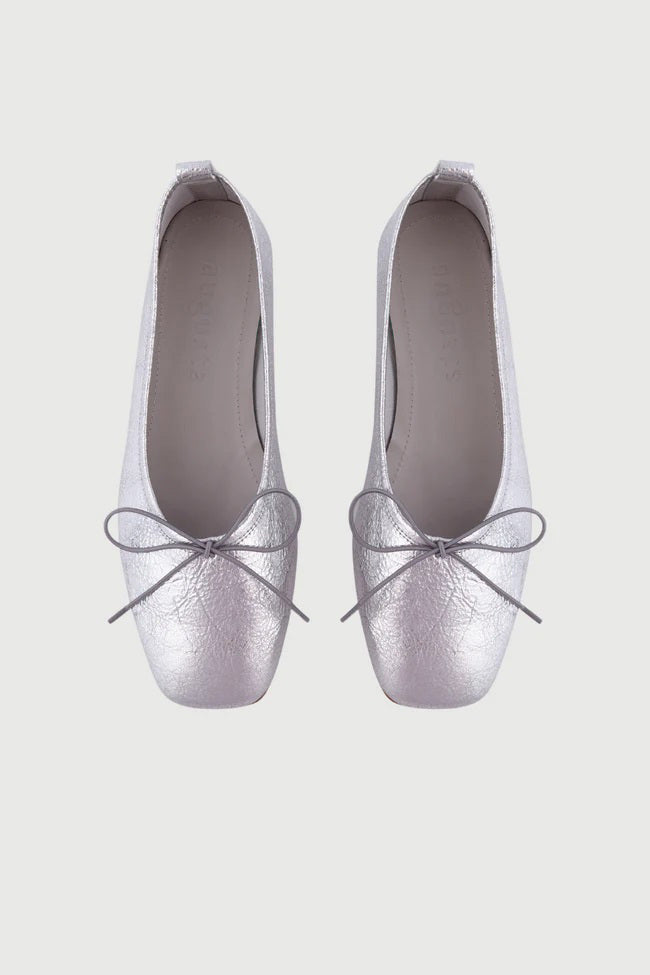 The Augusta - Sienna Ballet Flat by AUGUSTA is a single silver-colored shoe featuring a subtle metallic sheen and a simple bow on the front. It has a low brown sole and a pull tab at the back. Handcrafted in Spain from leather, this ballet flat is shown from the side against a light gray background.