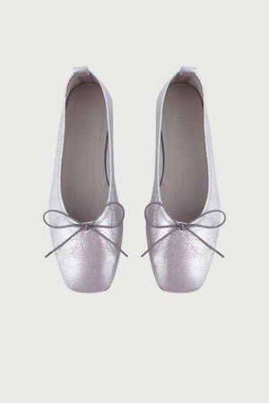 The Augusta - Sienna Ballet Flat by AUGUSTA is a single silver-colored shoe featuring a subtle metallic sheen and a simple bow on the front. It has a low brown sole and a pull tab at the back. Handcrafted in Spain from leather, this ballet flat is shown from the side against a light gray background.