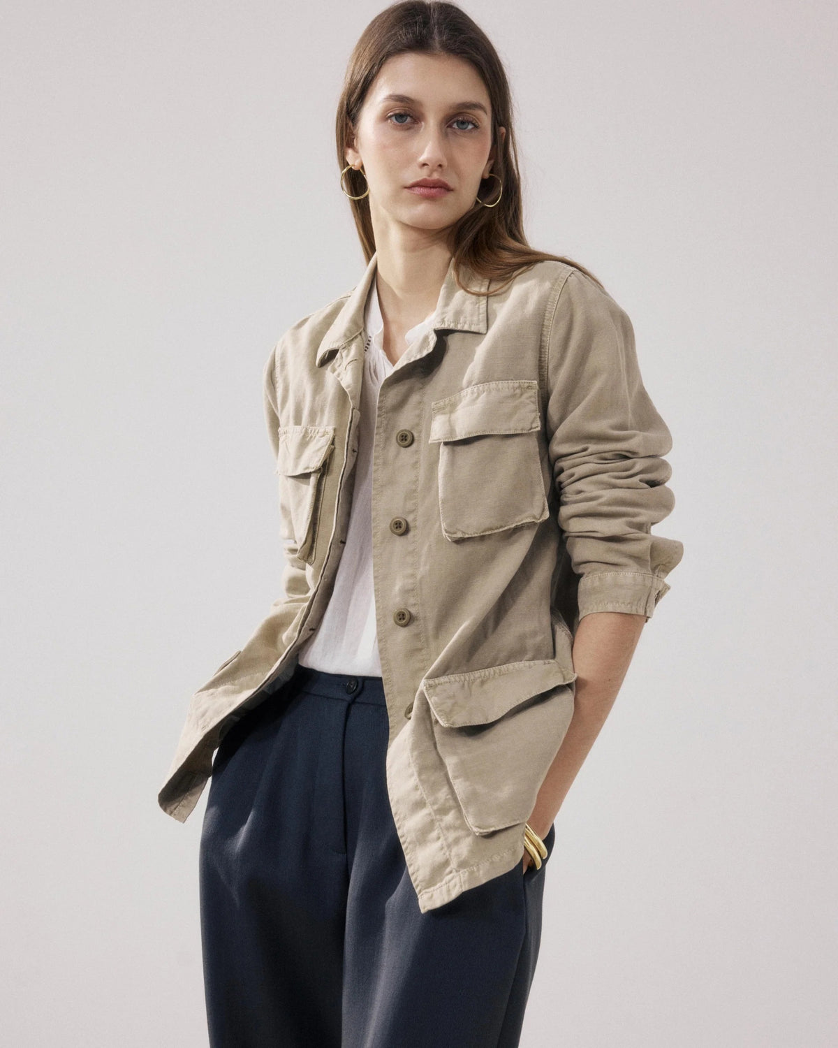 A woman stands indoors wearing the HARTFORD VEA Utility Jacket—a belted beige cotton-linen piece from HARTFORD—paired with dark loose pants, black sandals, and minimal jewelry. One arm is relaxed while the other is bent at the elbow. The background is plain and light.