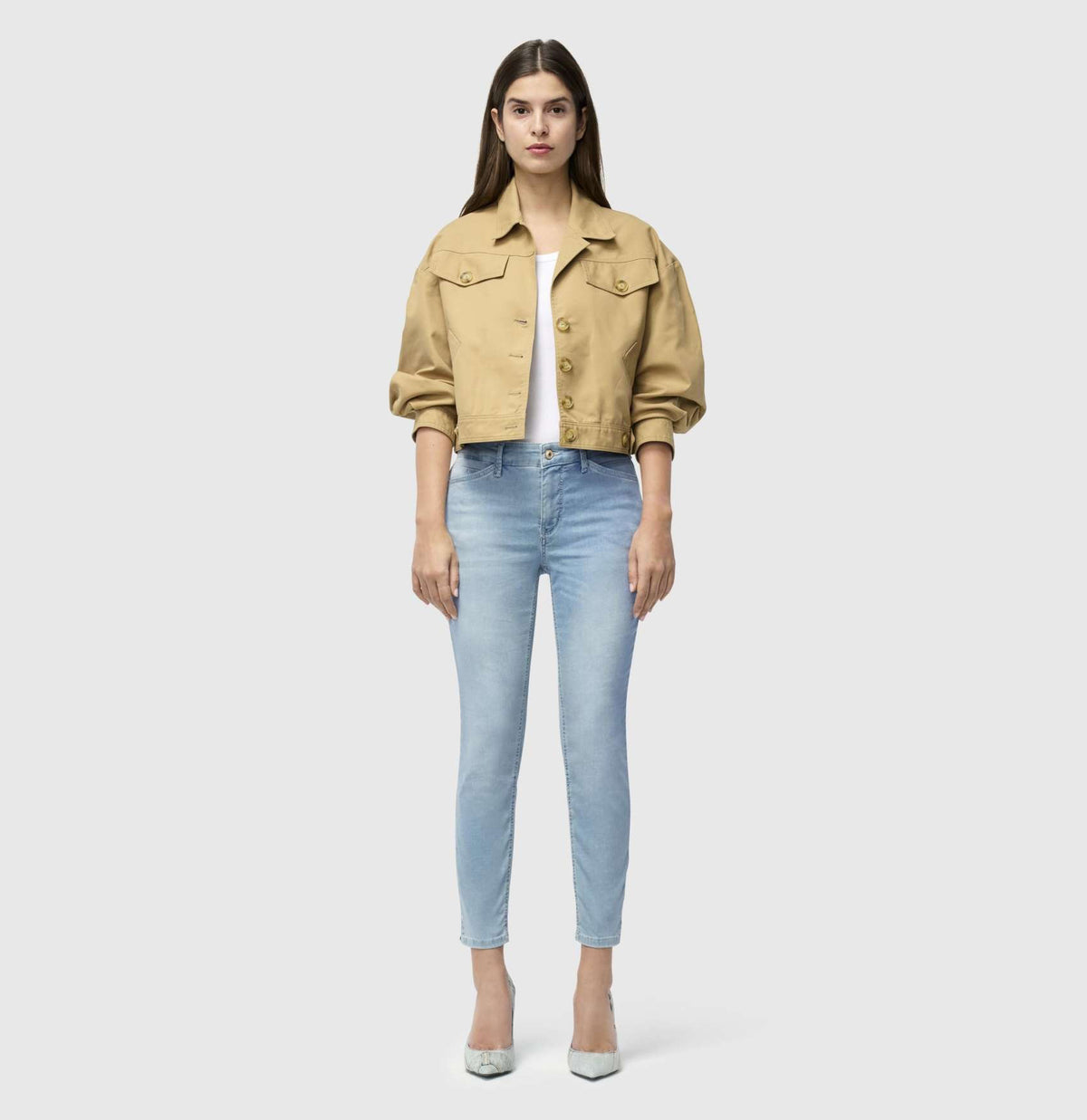 With long brown hair, a person stands against a plain backdrop wearing MAC&#39;s tan cropped jacket and MAC JEANS - DREAM SUMMER light blue slim jeans made from a comfortable modal blend. White heels complete the look as they gaze ahead with relaxed arms by their sides.