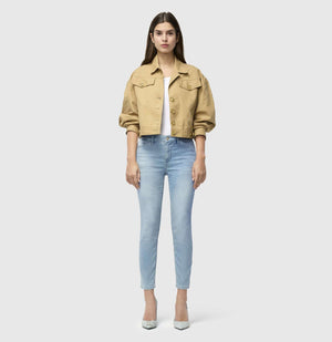 With long brown hair, a person stands against a plain backdrop wearing MAC's tan cropped jacket and MAC JEANS - DREAM SUMMER light blue slim jeans made from a comfortable modal blend. White heels complete the look as they gaze ahead with relaxed arms by their sides.