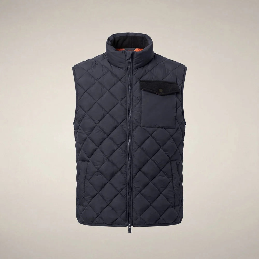 A person stands facing forward, wearing the SAVE THE DUCK - FABRICE Puffer Vest in Blue Black from SAVE THE DUCK, featuring recycled Plumtech insulation, layered over a black long-sleeve shirt and gray pants. They have a neutral facial expression and are posed against a plain white background.