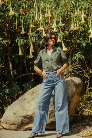 A woman in the KMJ - Southern Lover Shirt with a ruffle collar and blue jeans, featuring the iconic KMJ tuck, stands confidently beside a horse, holding its reins amidst green foliage. She's elegantly posed with jewelry, embodying Southern-inspired charm.