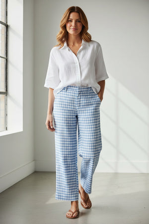 A person stands on a sandy beach wearing the FRANK & EILEEN WEXFORD Wide Leg Linen Pant in Blue Gingham, paired with a white shirt and beige sandals. The ocean is visible in the background.
