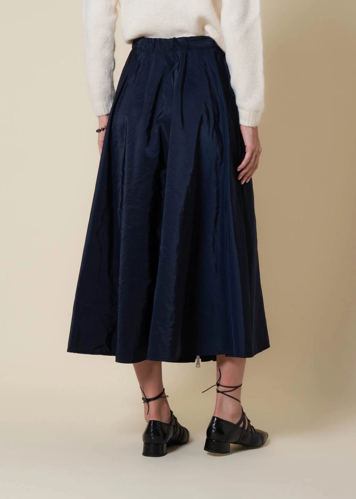 A woman in a cream sweater, scarf, and black heels stands by a large window, sunlight casting shadows on a beige wall, wearing the VICARIO CINQUE gazelle midi taffeta skirt with zipper in midnight blue.