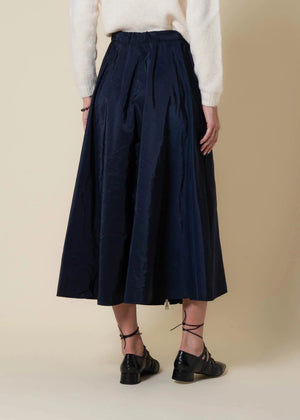 A woman in a cream sweater, scarf, and black heels stands by a large window, sunlight casting shadows on a beige wall, wearing the VICARIO CINQUE gazelle midi taffeta skirt with zipper in midnight blue.