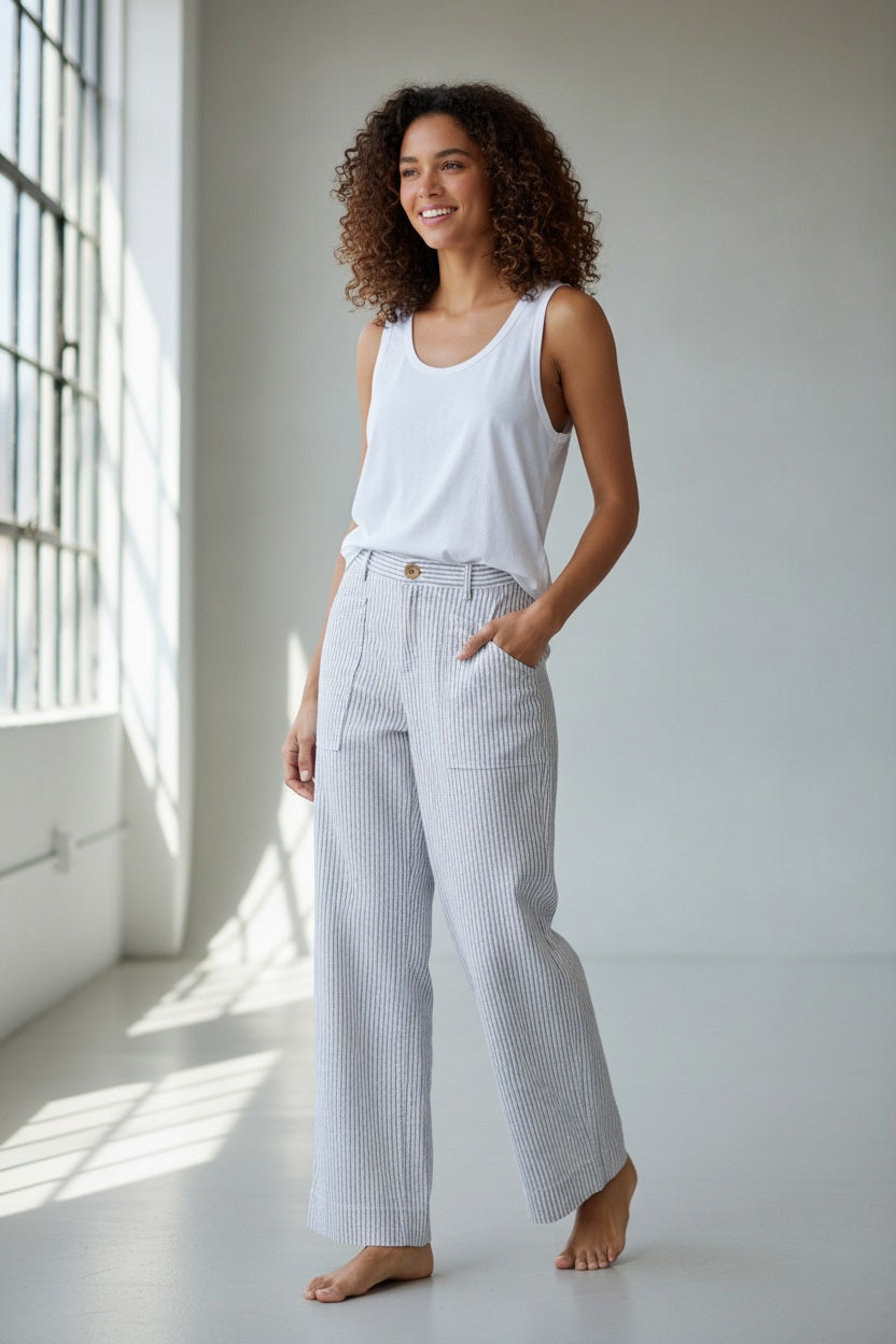 A person stands outdoors on grass, shown from the back, wearing the FRANK &amp; EILEEN Limerick Full Length Wide Leg Carpenter Pant in Italian Performance Linen Navy and White Stripe and a white top, with one hand resting on their hip. Shrubs and a stone border are in the background.