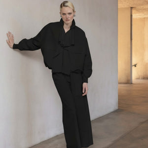 A woman faces forward against a plain background, wearing a black top, blue jeans, white sneakers, and the NUDE Lightweight Bomber Jacket with Ruffle by NUDE draped over her shoulders. Her dark hair is loose and she looks relaxed.