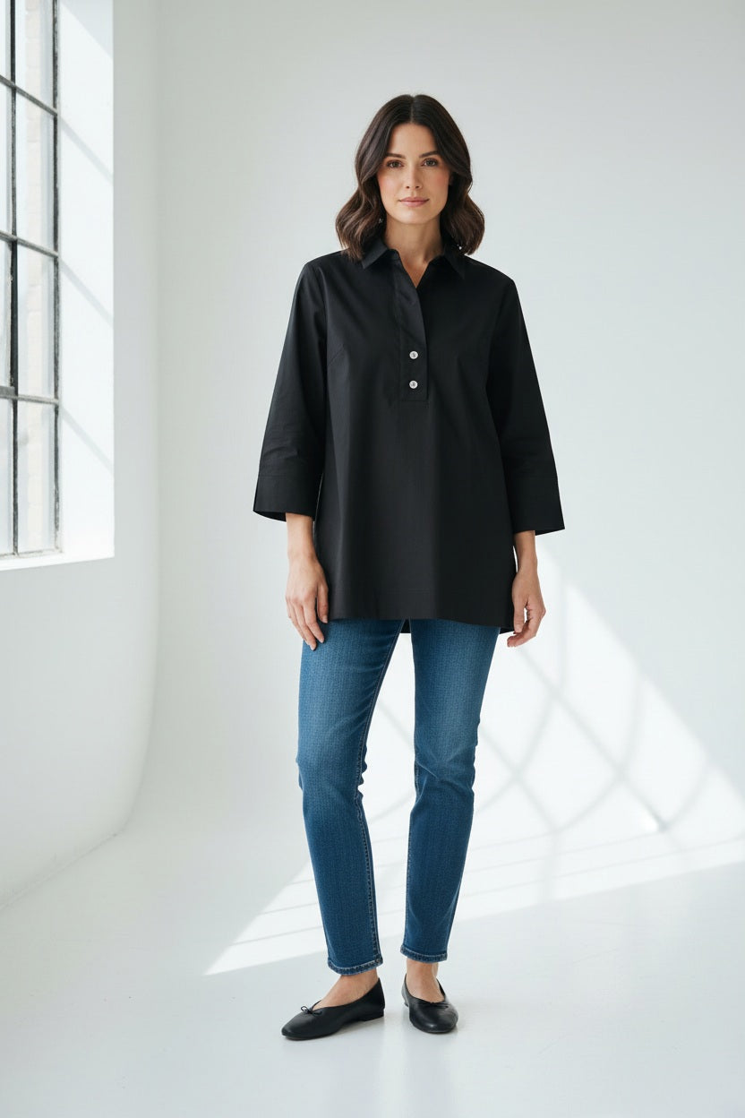 A woman wearing the HINSON WU Aileen Tunic Shirt—a navy blue button-up with three-quarter sleeves and tunic-length—paired with dark gray pants, stands against a white wall, facing slightly to the side.