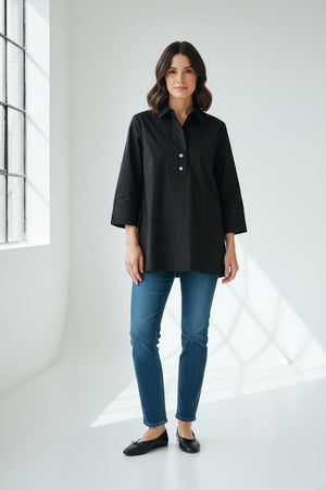 A woman wearing the HINSON WU Aileen Tunic Shirt—a navy blue button-up with three-quarter sleeves and tunic-length—paired with dark gray pants, stands against a white wall, facing slightly to the side.