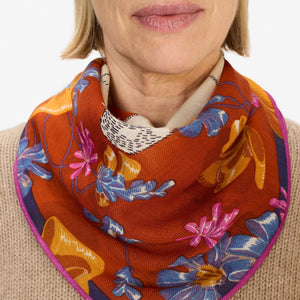 A woman with blonde hair, wearing a tan sweater and the INOUI TOUTOU DOG SCARF IN ORANGE, stands facing the camera against a plain white background.