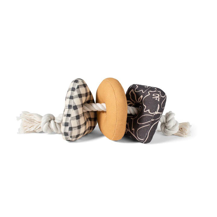 Introducing the PET SHOP BY FRINGE STUDIO - ROPED IN CANVAS DOG TOY: This interactive toy features a sturdy white rope threaded through three engaging shapes—a black-and-white checkered triangle, a yellow donut-shaped ring, and a square adorned with black and white floral designs. Made from durable cotton canvas for lasting play, the securely knotted rope ends ensure added safety.