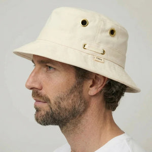 A bearded man in a white shirt wears the TILLEY - ICONIC T1 BUCKET HAT IN NATURAL with UPF 50+ protection and metal eyelets, looking directly at the camera against a plain background.
