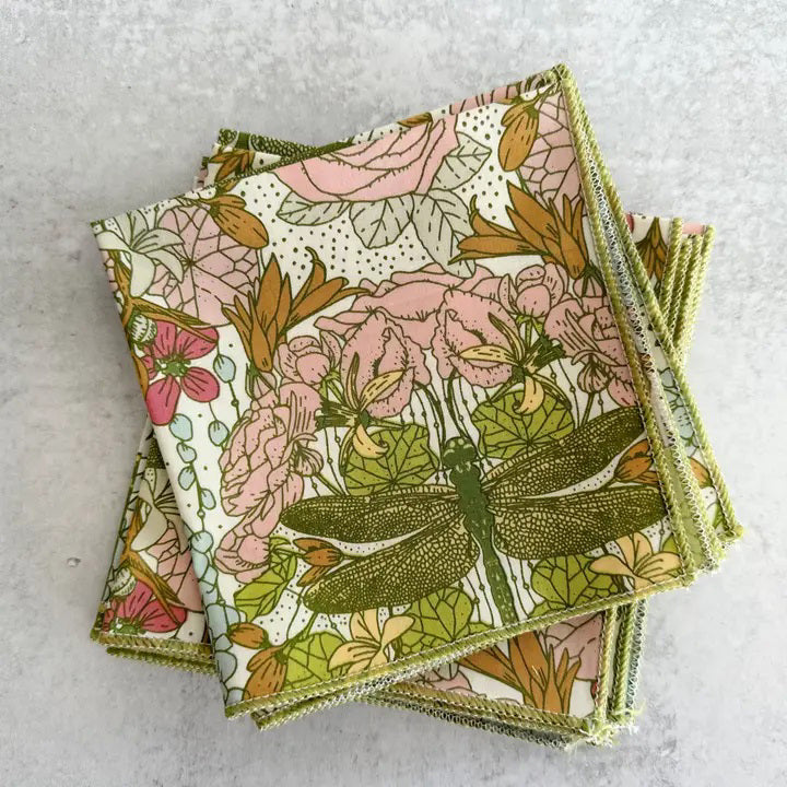 A neat stack of DRAGONFLY GARDEN COCKTAIL NAPKINS by DOT AND ARMY, made from 100% cotton. These super soft napkins feature a vibrant floral print with pink and white flowers, green leaves, and a large green dragonfly in the center, all set on a light gray surface.