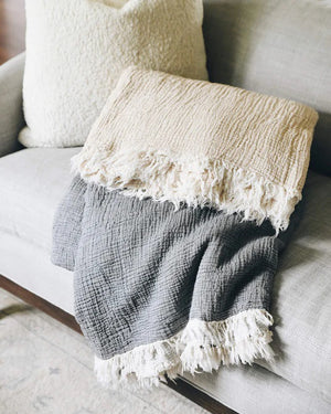 The MERSEA CLOUD GAUZE BLANKET, shown folded on a light background, is a soft beige cotton throw with a textured surface and fringed edges.