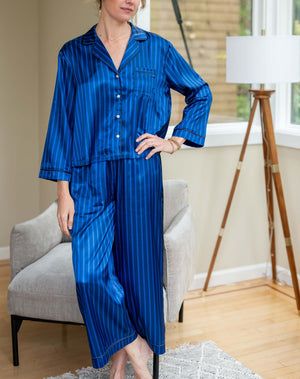 The MERSEA - Carousel Stripe Satin Sailor Pajama Set in Blue by MERSEA, featuring elegant blue and black stripes with a button-up top and matching pants, embodies luxury sleepwear and is neatly folded on a light blue background.