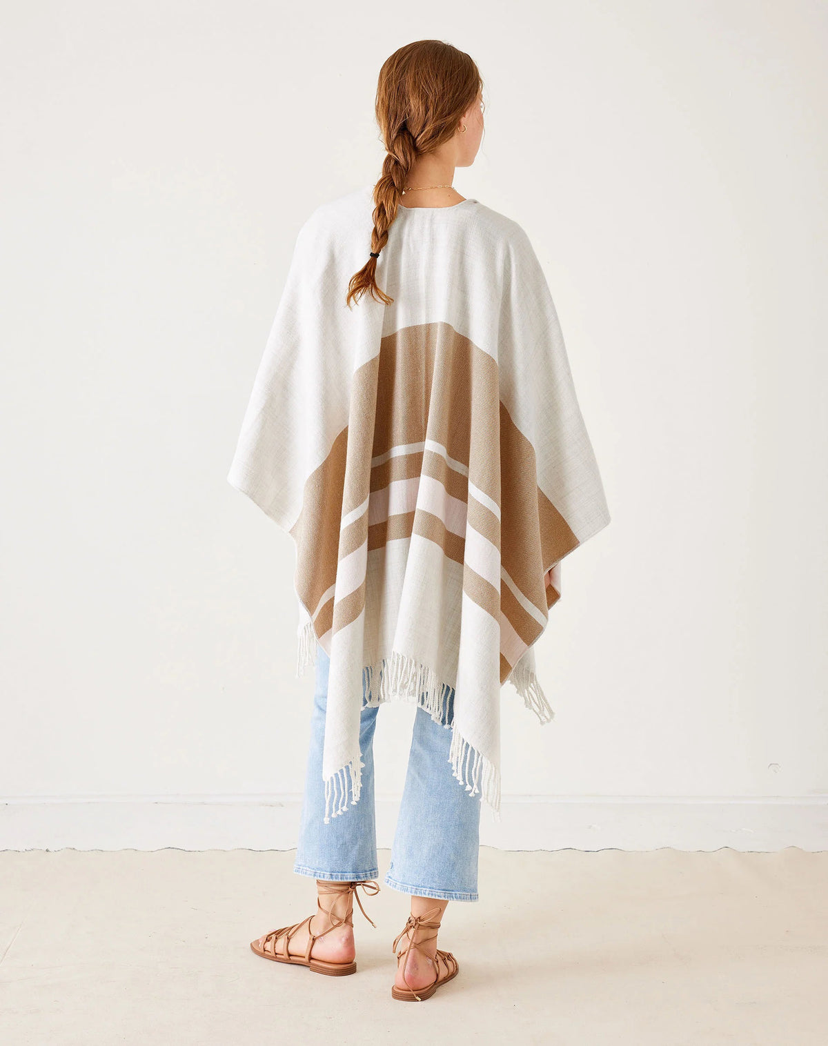 A woman stands against a white wall, wearing the MERSEA - CLASSIC TRAVEL WRAP IN DUNE STRIPE over a pale pink blouse, light blue jeans, and tan sandals. She faces the camera with a neutral expression.