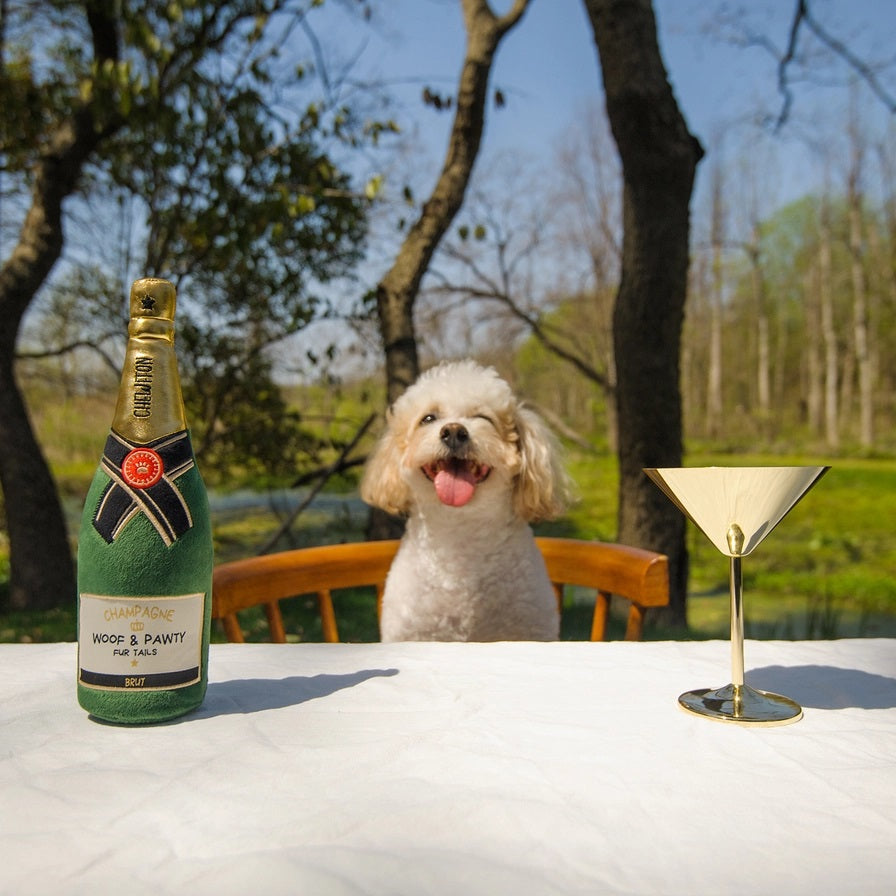 A brown and white spotted dog stands on a light background, holding the CHEWFFON PLUSH CHAMPAGNE BOTTLE SQUEAKY DOG TOY in its mouth.