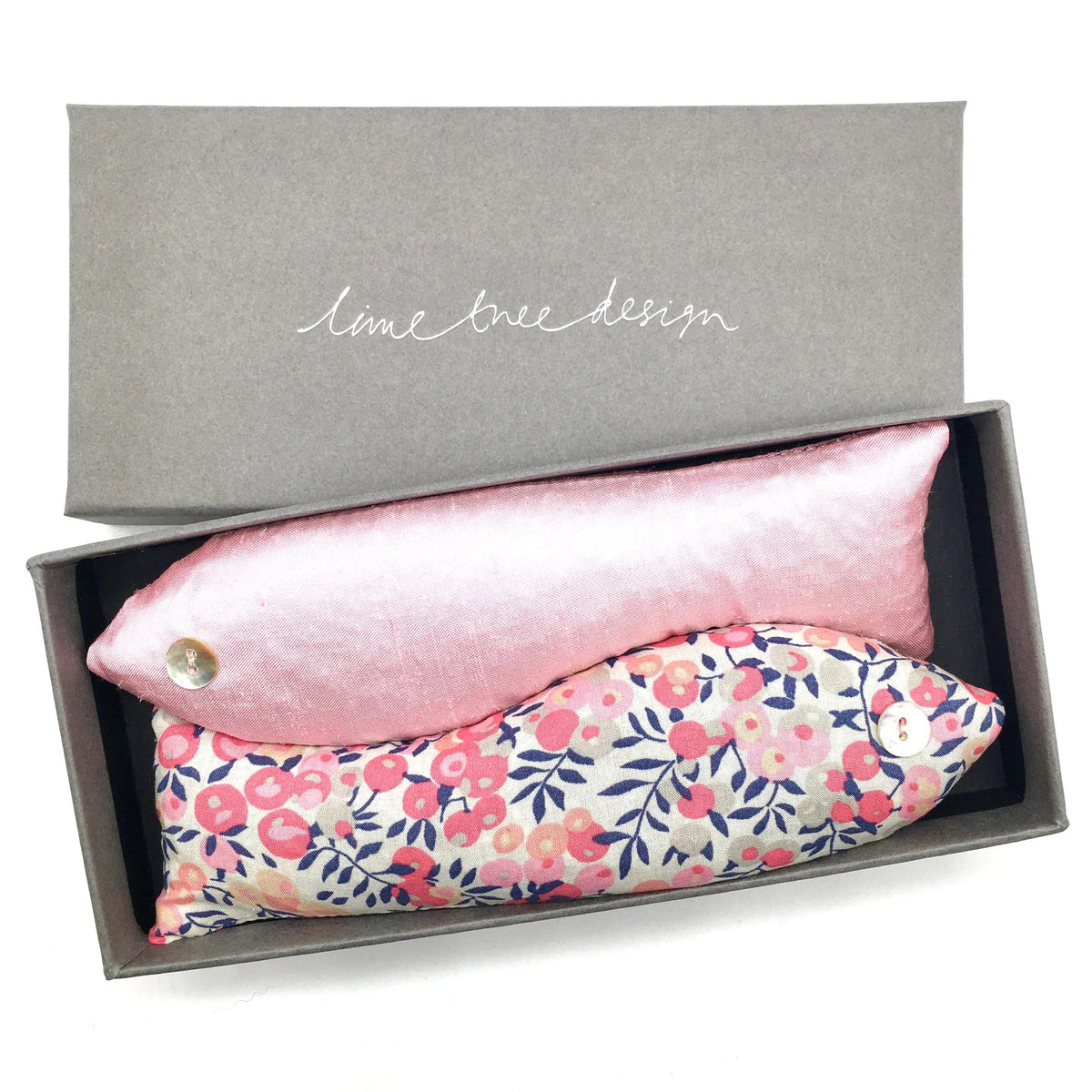 A rectangular grey box from the LIME TREE COLLECTION LTD, featuring &quot;LIME TREE DESIGN&quot; on the lid, contains two handmade lavender sardines. One fish is pink with a button eye, while the other is crafted from Liberty Tana Lawn fabric and showcases a floral pattern in pink, blue, and yellow. The box is open with the lid placed beside it. This charming set is named SACHET LAVENDER FISH.