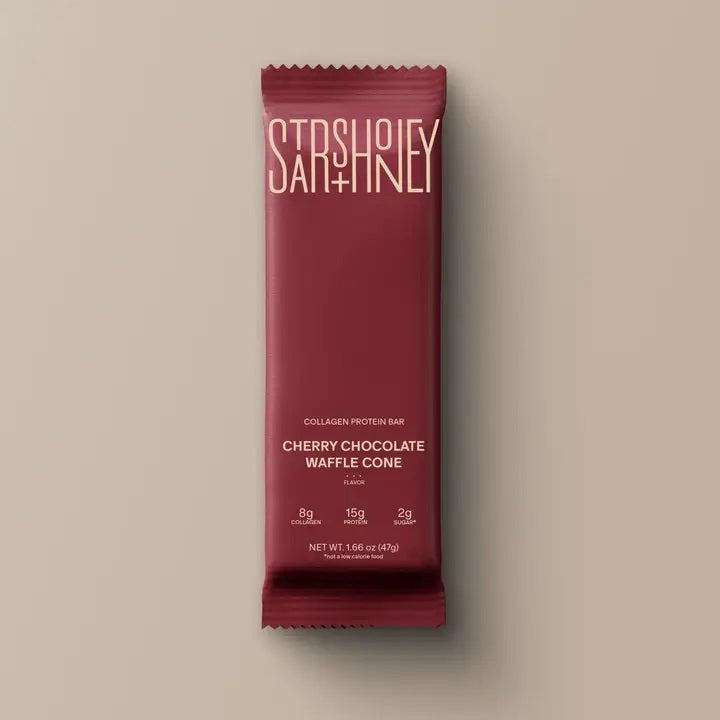 A maroon protein bar wrapper labeled “STARS+HONEY” displays the flavor “CHERRY CHOCOLATE WAFFLE CONE” with nutrition highlights: 8g collagen, 15g protein, and 2g sugar, all set against a beige background. This indulgent treat puts the rich chocolate and tart cherry combination front and center.