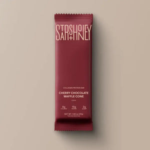 A maroon protein bar wrapper labeled “STARS+HONEY” displays the flavor “CHERRY CHOCOLATE WAFFLE CONE” with nutrition highlights: 8g collagen, 15g protein, and 2g sugar, all set against a beige background. This indulgent treat puts the rich chocolate and tart cherry combination front and center.