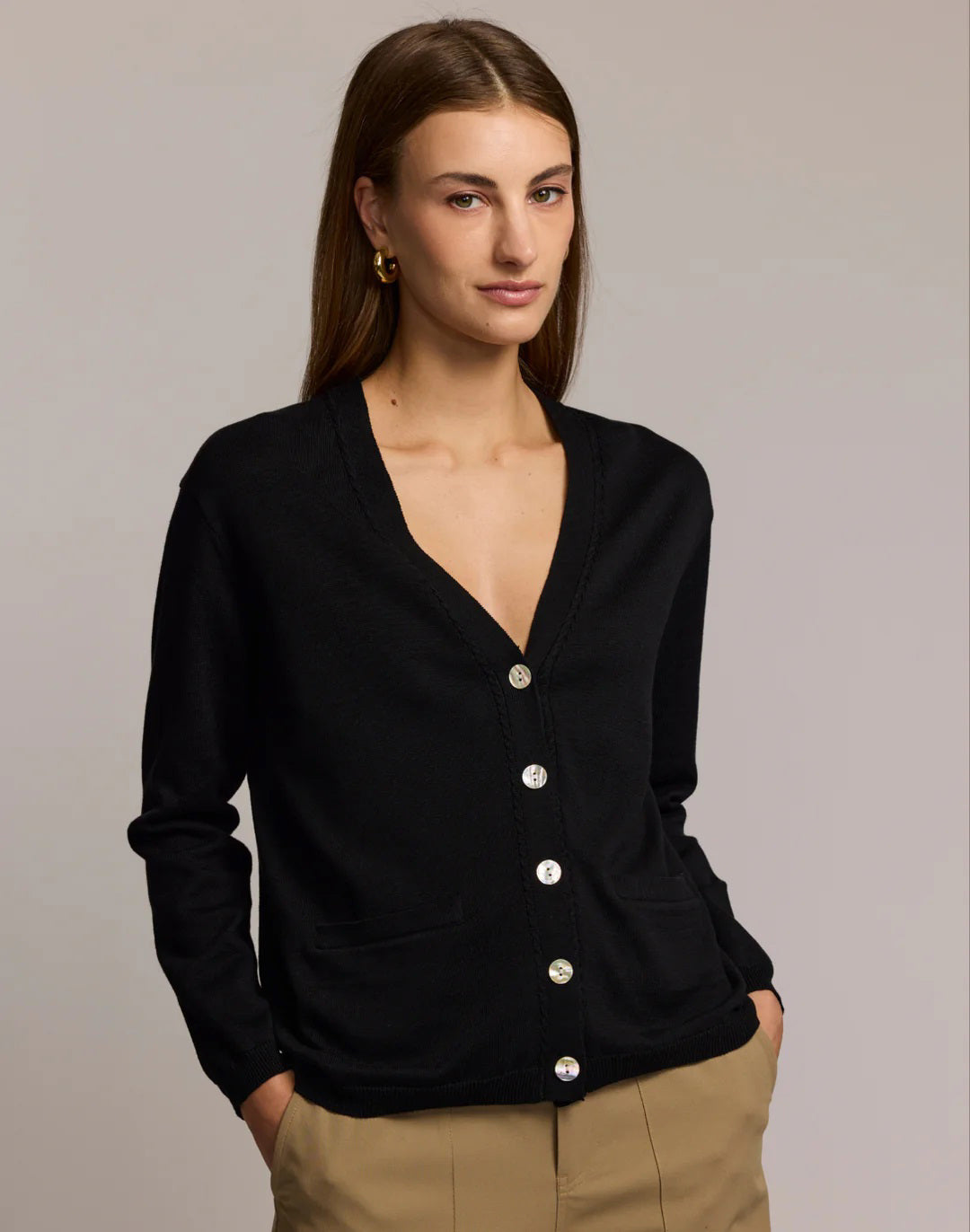 A woman with straight brown hair wears the HINSON WU CASSIE Long Sleeve Cotton Silk Cardigan in camel, styled with black pants. She poses confidently, one hand to her chin, against a neutral backdrop.