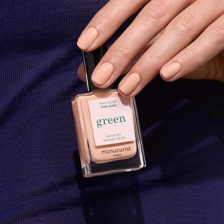 A bottle of MANUCURIST - GREEN™ NAIL POLISH IN PINK SAND by MANUCURIST INC, presented in a rectangular glass bottle with a black cap and a label displaying “green,” along with the phrases “quick dry” and “intense color.”.